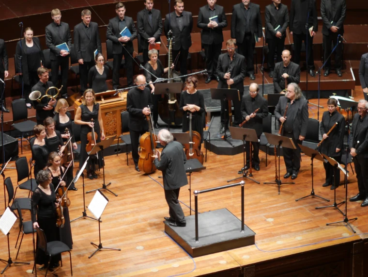 A choir and period-instrument orchestra in black stand on the wooden stage of the Usher Hall around the conductor's podium at the close of a performance (2014). The Edinburgh International Festival, Usher Hall, Edinburgh, Scotland, in the season calendar of The Mansa Court
