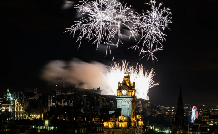 Fireworks burst over Edinburgh Castle at night, with the lit clock tower of the Balmoral Hotel and the spire of the Scott Monument in the foreground (2015). The Edinburgh International Festival, Edinburgh, Scotland, in the season calendar of The Mansa Court