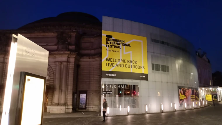 The Usher Hall at dusk under a deep blue sky, its curved glass extension hung with a yellow Edinburgh International Festival banner reading "7–29 August, Welcome back live and outdoors" (2021). The Edinburgh International Festival, Usher Hall, Edinburgh, Scotland, in the season calendar of The Mansa Court