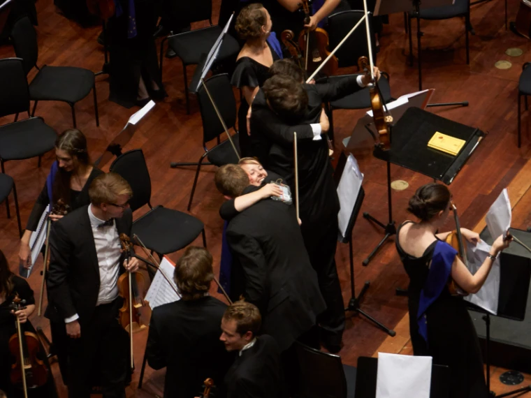 Seen from above, young orchestra musicians in black evening dress embrace among music stands and chairs on stage after a concert (2015). The Edinburgh International Festival, Usher Hall, Edinburgh, Scotland, in the season calendar of The Mansa Court