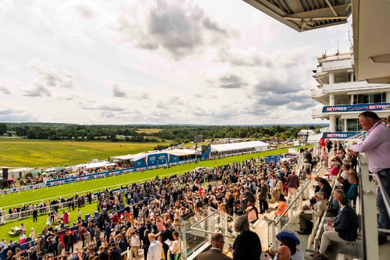 Racegoers crowd the lawns and the rail below the grandstand, the Downs rolling away beyond (2025). The Derby at Epsom Downs, Surrey, in the season calendar of The Mansa Court