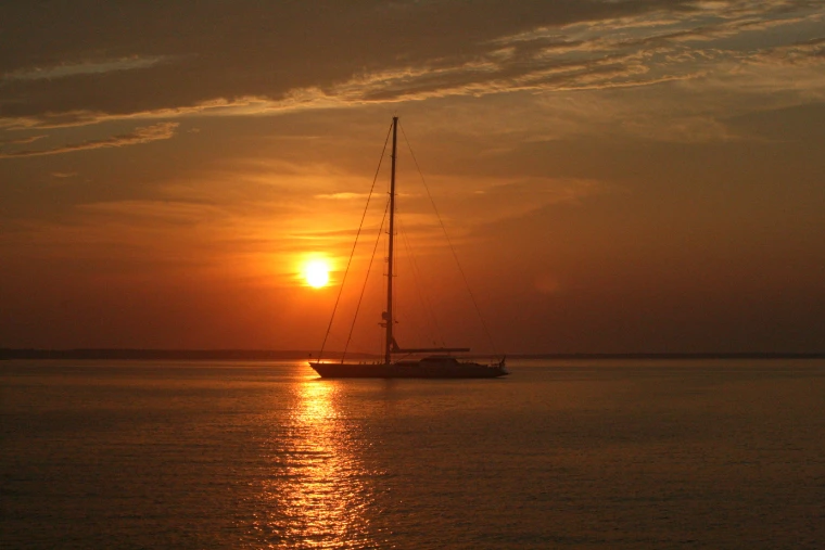 A large sailing yacht with sails furled lies silhouetted on calm water as the sun sets behind her mast (2011).
