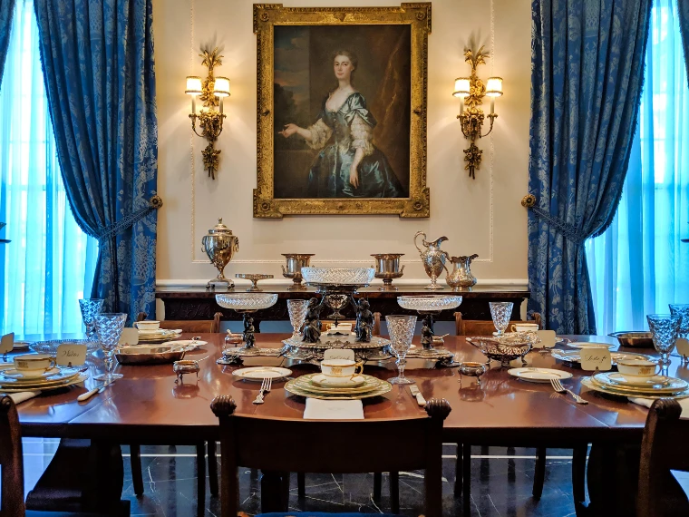 A mahogany dining table laid with china, crystal glasses and silver beneath a gilt-framed portrait, between blue damask curtains and a sideboard of silver (2019).