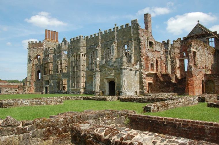 The roofless stone and brick walls of the Cowdray ruins rise above low foundations and green lawn under a blue sky (2013).