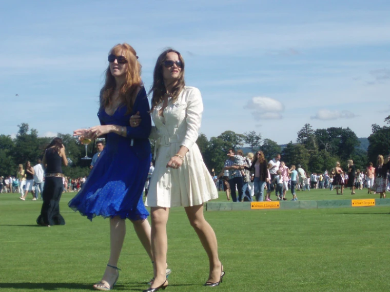 Two women in a blue dress and a cream coat dress walk arm in arm across the polo field among other spectators (2007).