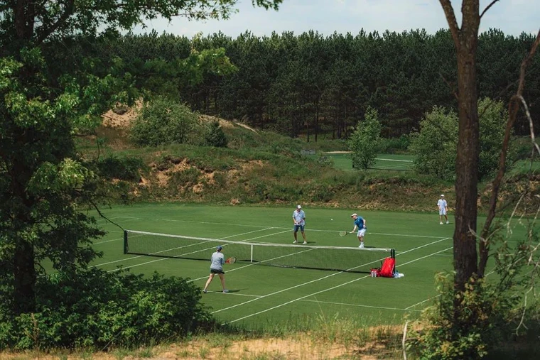 Four players at doubles on a grass court cut into a wooded hollow, pines on the ridge behind, for The Grass Court Tennis Tournament, Southampton, New York, an afternoon in the season calendar of The Mansa Court
