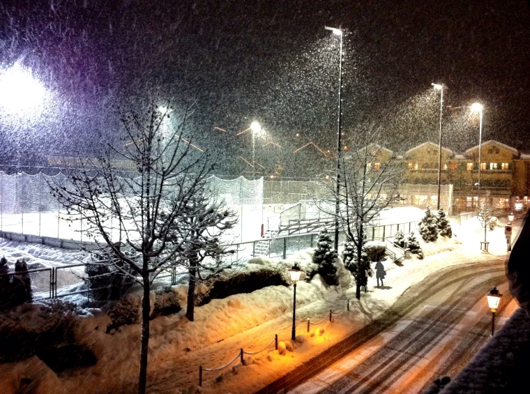 Heavy snow falling at night under streetlamps over a snowbound road and the floodlit ice rink, chalets glowing in the background (2012).