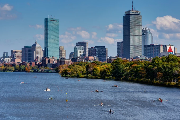 Small boats and crews spread across the Charles River beneath the Boston skyline and autumn trees on a sunny day (2014).