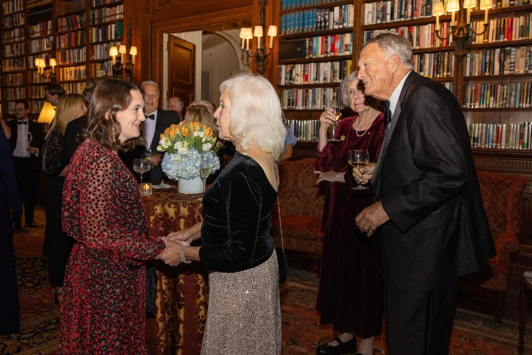 Guests in evening dress greet one another among drinks in a wood-panelled library lined with books and lit by sconces (2025).