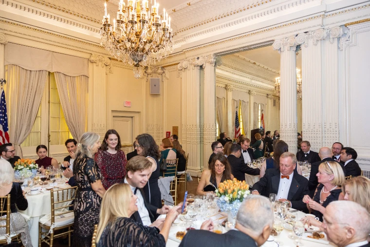 Guests in black tie and evening dress at round tables set with orange tulips in a white-and-gold ballroom under a crystal chandelier, Dutch and American flags beyond (2025).