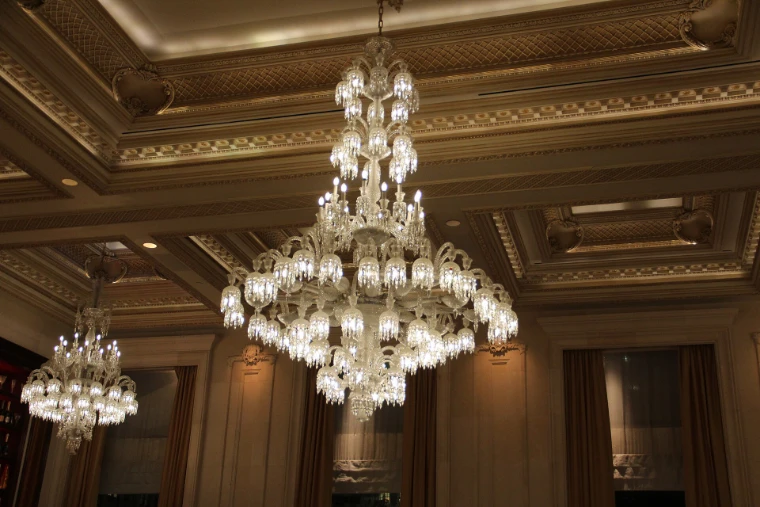 Tall crystal chandeliers hanging beneath a gilded, coffered ceiling in a room of the Plaza Hotel (2017).