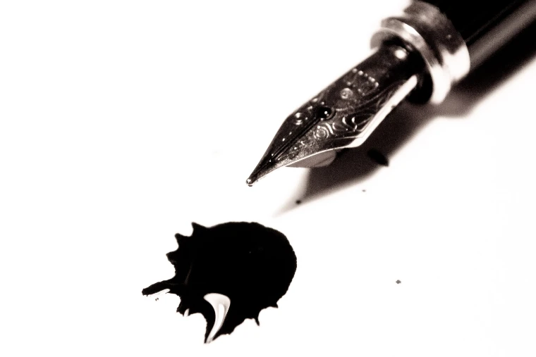 The nib of a fountain pen above a fresh blot of black ink on white paper, in black and white (2013).