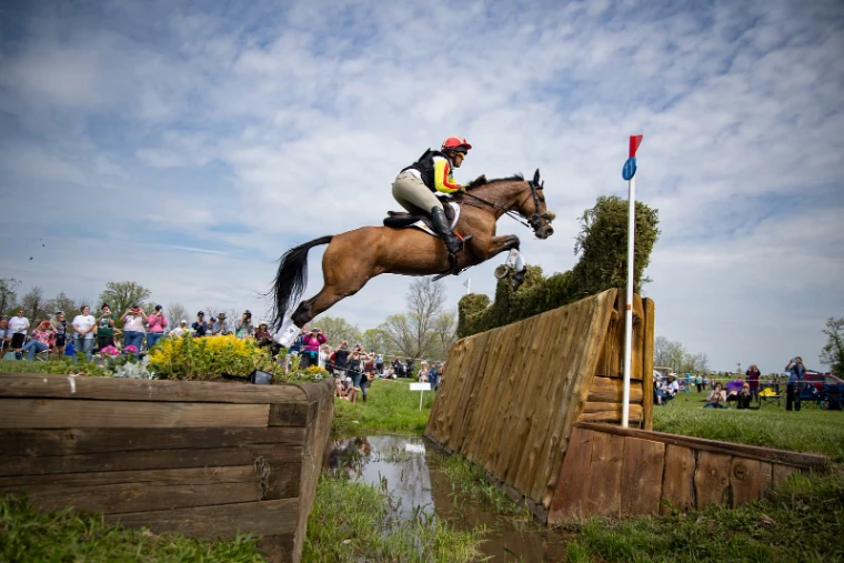 A bay horse and rider in red and yellow flying a brush fence over the ditch on the cross-country course at the Kentucky Three-Day Event, Lexington, a late-April engagement in the season calendar of The Mansa Court