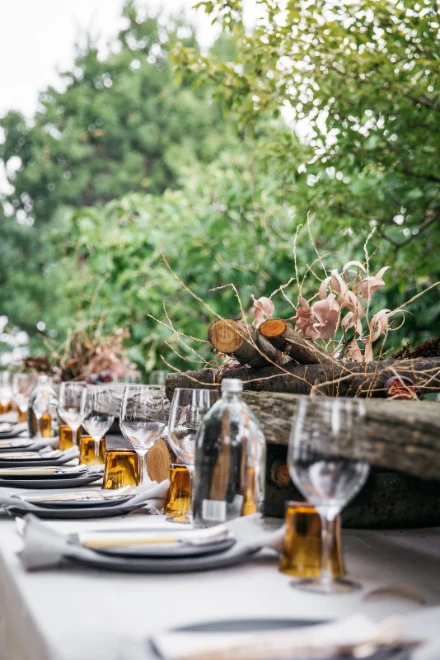 A long table laid with white plates, glasses and bottles beneath greenery and flowers, for The Long Table Sunday Lunches, New York, June Sundays in the season calendar of The Mansa Court