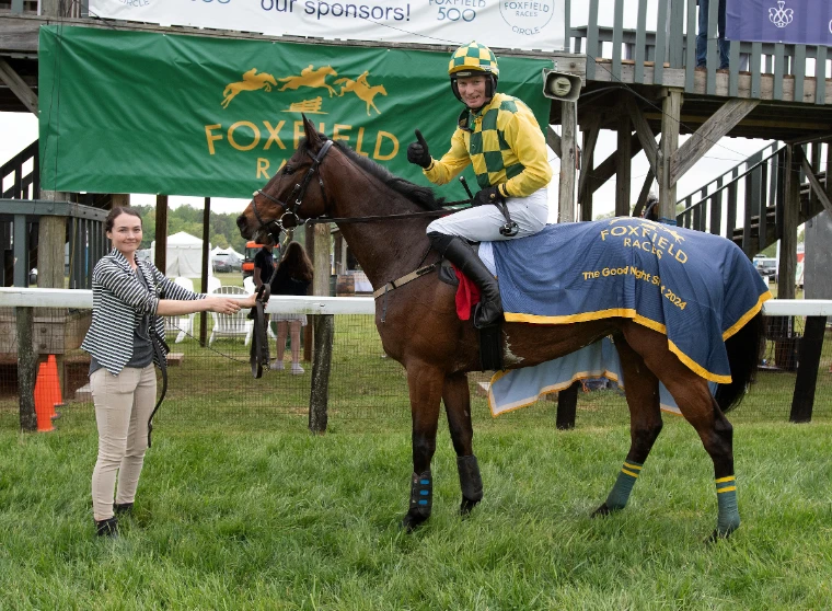 A jockey in green and yellow checks giving a thumbs-up from the saddle of a winning chaser in the enclosure at Foxfield, on the spring timber circuit that leads to The Maryland Hunt Cup, Glyndon, a late-April engagement in the season calendar of The Mansa Court