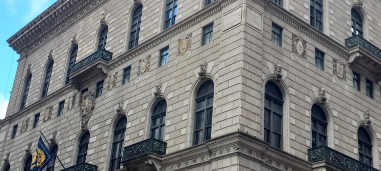The arched windows, iron balconies and carved college crests of the University Club’s façade, with its blue banners hanging (2024).