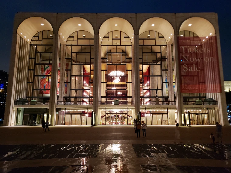 The five tall arches of the Metropolitan Opera House glow from within at night, chandeliers and red walls visible through the glass, above a rain-wet plaza with a few people walking (2018).