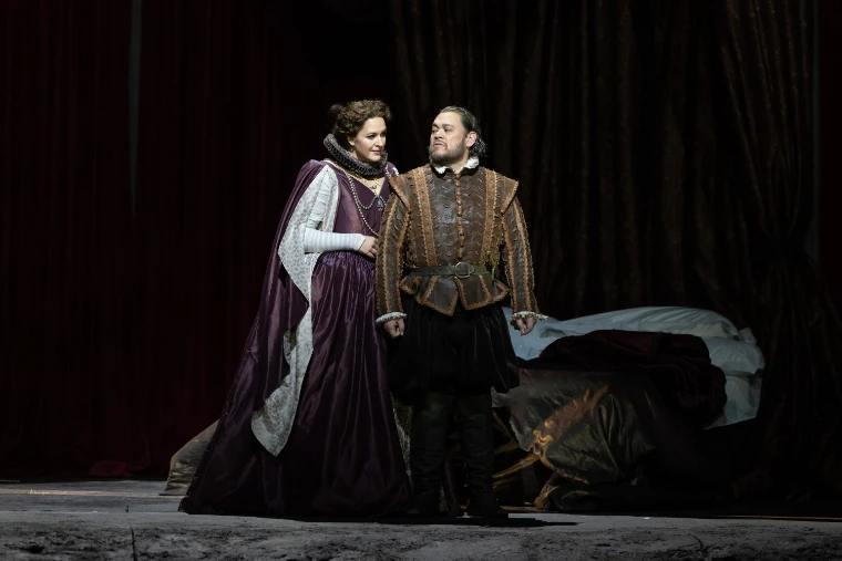 Lise Davidsen as Lady Macbeth, in a purple gown and ruff, stands beside Quinn Kelsey as Macbeth before a dark curtain and an unmade bed in the Met’s new production of Verdi’s Macbeth (2026).