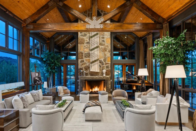 The great room of a timber-framed mountain house, exposed trusses, a lit fire in a tall stone chimney, pale sofas and tall windows onto the trees, for The Mountain Cabin, Aspen, Colorado in the season calendar of The Mansa Court