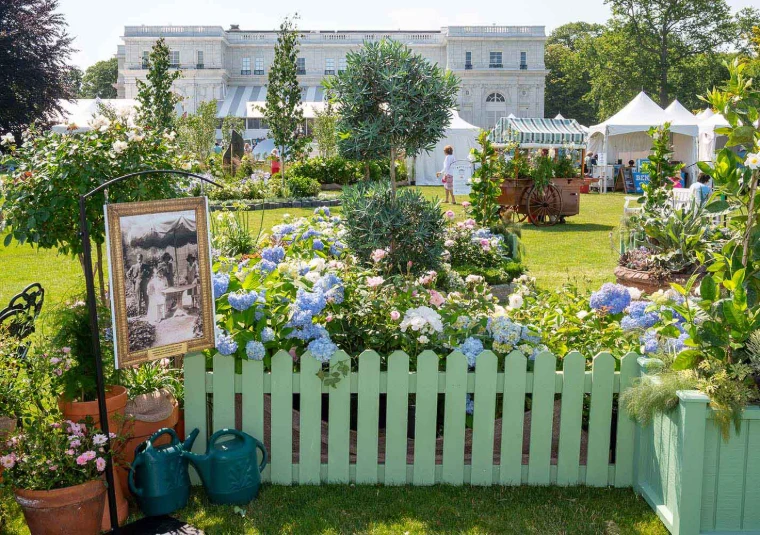 Hydrangeas behind a green picket fence on the lawn, with Rosecliff and the show tents beyond. The Newport Flower Show at Rosecliff, Newport, in the season calendar of The Mansa Court