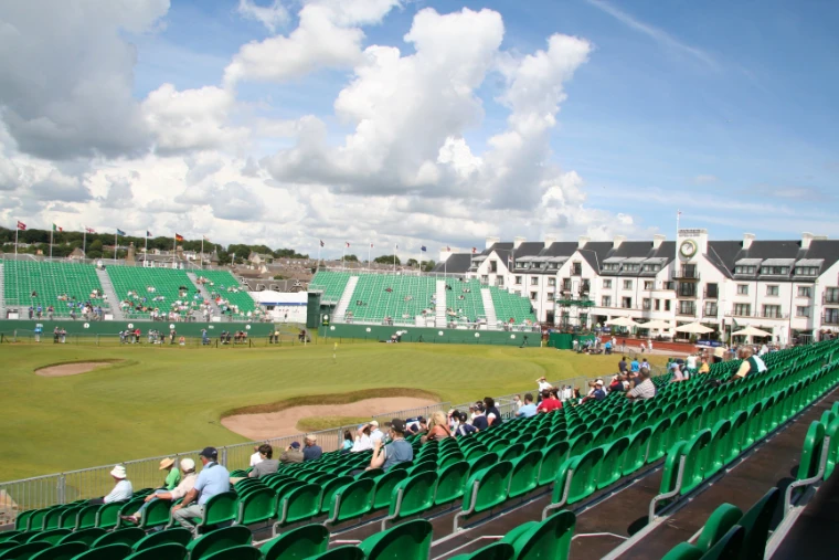 Tiers of green grandstand seats ring the 18th green and its bunkers, with flags flying and a long white hotel beyond under a bright sky of white clouds (2007).