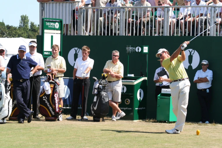 A golfer in a yellow shirt follows through on his tee shot from the first tee as players, caddies and officials watch beneath a grandstand full of spectators (2006).