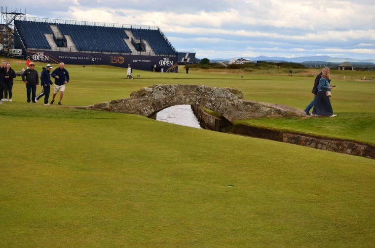 The old stone Swilken Bridge crosses the burn on the Old Course, with walkers on the fairway and a large grandstand bearing The Open signage behind (2022).