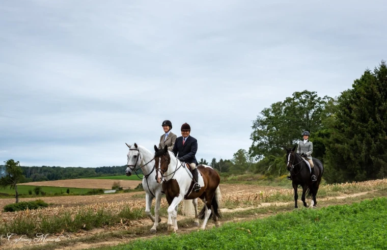 Riders, most in tweed coats, hacking along a grass track between fields at the edge of a wood.