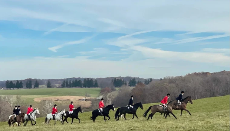Mounted members in scarlet and black coats riding in a line across a hillside, with open fields and autumn woods beyond.