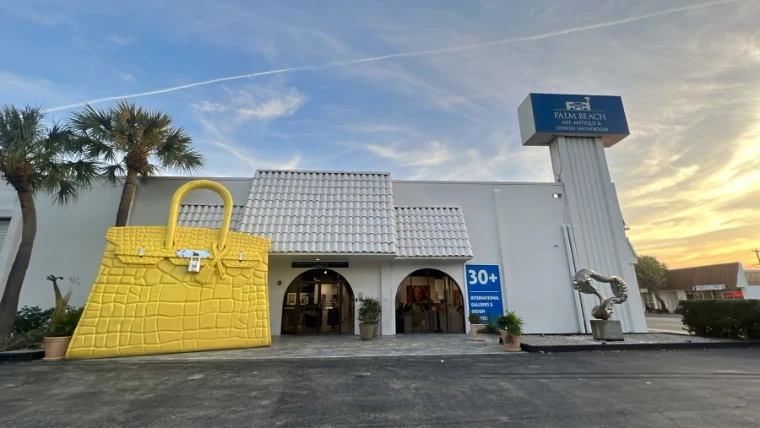 The Palm Beach Art, Antique and Design Showroom at sunset, a giant yellow handbag sculpture beside its arched entrance, at The Palm Beach Show, West Palm Beach, an engagement in the season calendar of The Mansa Court
