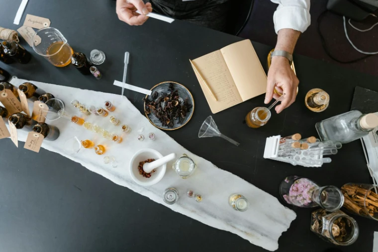 A dark table seen from above, laid with a white cloth, small bottles and vials, bowls of dried petals and resin and an open notebook, hands at work, for The Perfumer’s Evening, New York in the season calendar of The Mansa Court