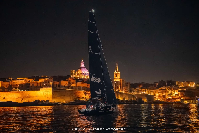 At night a racing yacht with dark sails comes in on water lit gold by the floodlit walls of Valletta, with a pink-lit dome and a spire behind (2025).