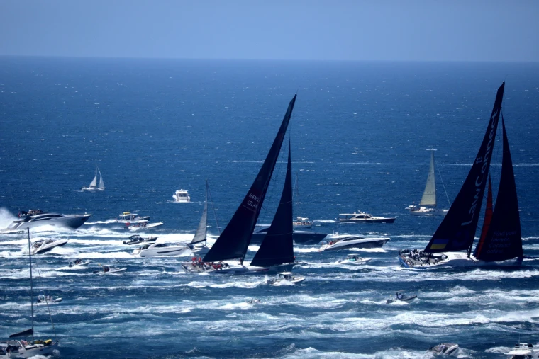 Two pairs of racing yachts under dark sails cross deep blue water churned white by the wakes of a crowd of spectator boats (2022).