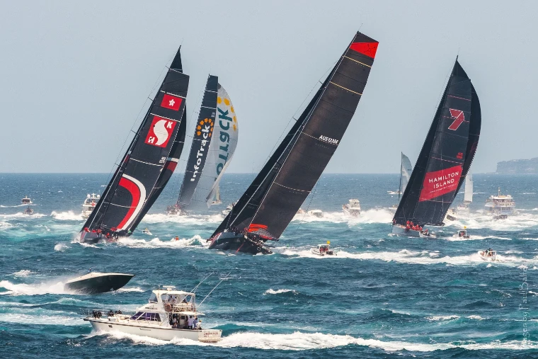 Four maxi yachts with black sails heel hard through choppy water at Sydney Heads after the Boxing Day start, spectator boats racing alongside (2019).