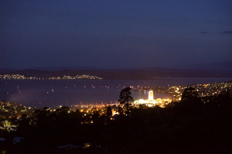 Hobart’s waterfront lit at nightfall beside the dark River Derwent, with the lights of boats on the water and hills beyond (2015).