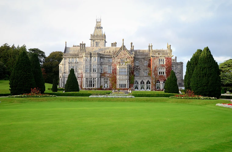 The grey stone Tudor Revival front of Adare Manor, with red creeper on its walls, clipped yews and flower beds, seen across a wide lawn (2013).