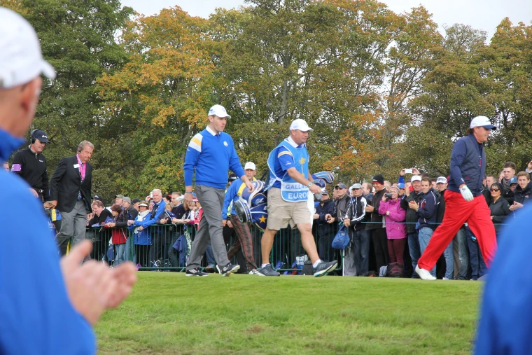 Two golfers, one in blue and one in red trousers, walk onto a tee with a caddie in a European bib, a crowd behind the barriers and autumn trees beyond (2014).