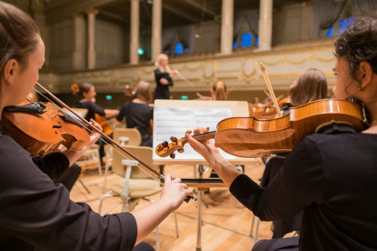 Young violinists playing from their music stands in a gilded hall, a conductor in the distance, for The Salon de Musique, New York in the season calendar of The Mansa Court