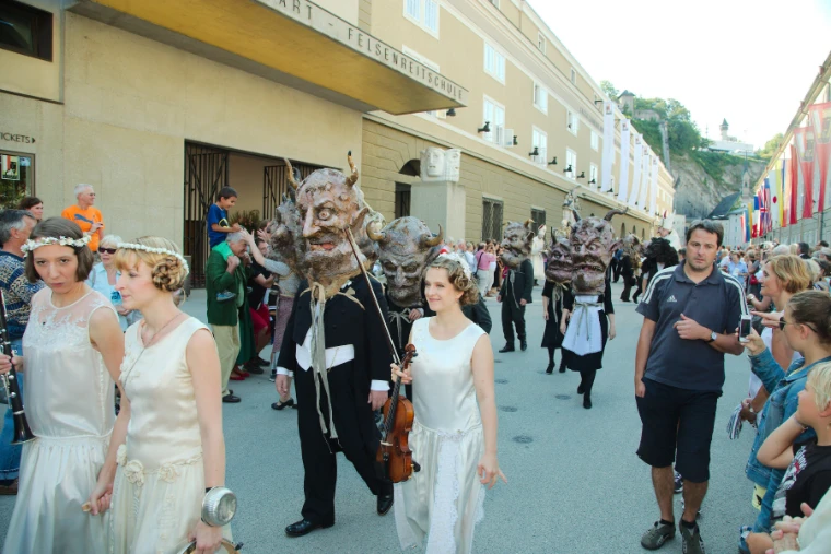 Costumed performers in white dresses and masked figures parade through a crowded street beside the festival halls, with the fortress hill behind (2013).