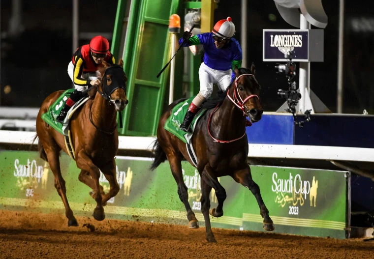 A jockey in blue and green silks raising his whip as he crosses the line ahead of the field at The Saudi Cup, Riyadh, an engagement in the season calendar of The Mansa Court