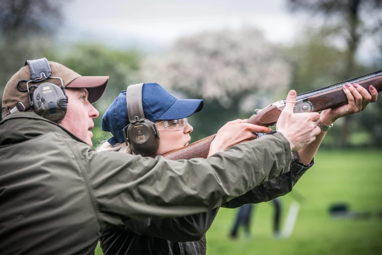 A shooting instructor in a cap and ear defenders steadying a woman’s over-and-under shotgun as she mounts it, blossom trees behind, for The Shooting School, Millbrook, New York, mid April in the season calendar of The Mansa Court