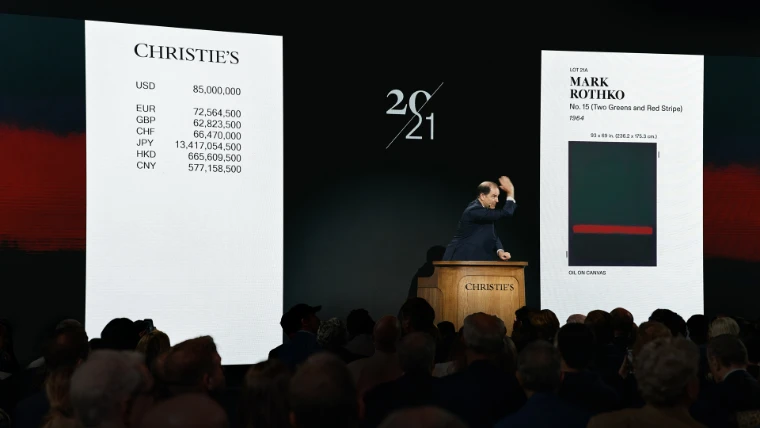 An auctioneer at the wooden Christie's rostrum raises his hand between two large screens, one showing bids in several currencies and the other a Mark Rothko painting, above a packed saleroom in silhouette (2026). The Spring Auctions, Christie's, Rockefeller Center, New York, in the season calendar of The Mansa Court