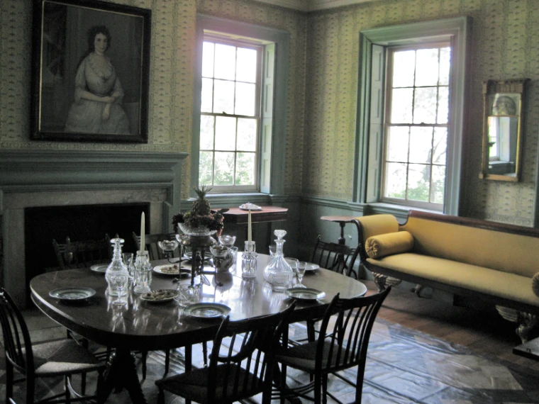 A period dining table laid with crystal decanters, candlesticks, plates and a pineapple centrepiece in a papered room with tall windows and a yellow sofa (2010). Morris-Jumel Mansion, Washington Heights, Manhattan, New York, shown for The Summer Table in the season calendar of The Mansa Court