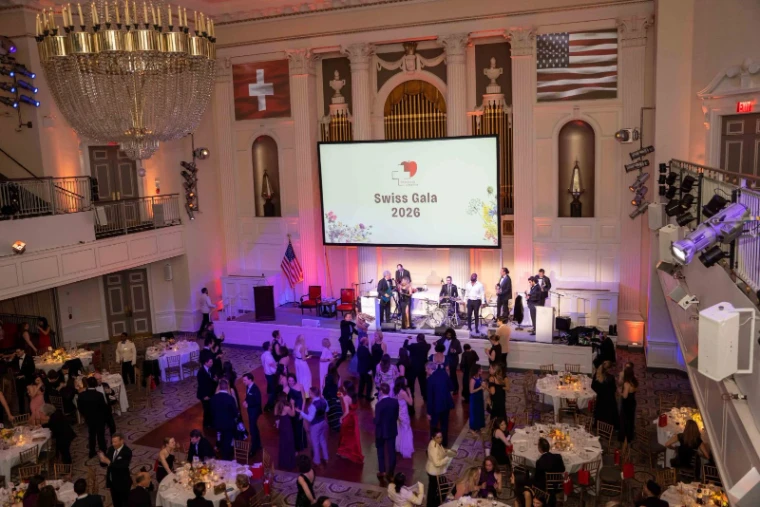 The ballroom seen from the balcony at The Swiss Ball, guests dancing before the band beneath the Swiss and American flags, the March black-tie evening in New York in the season calendar of The Mansa Court