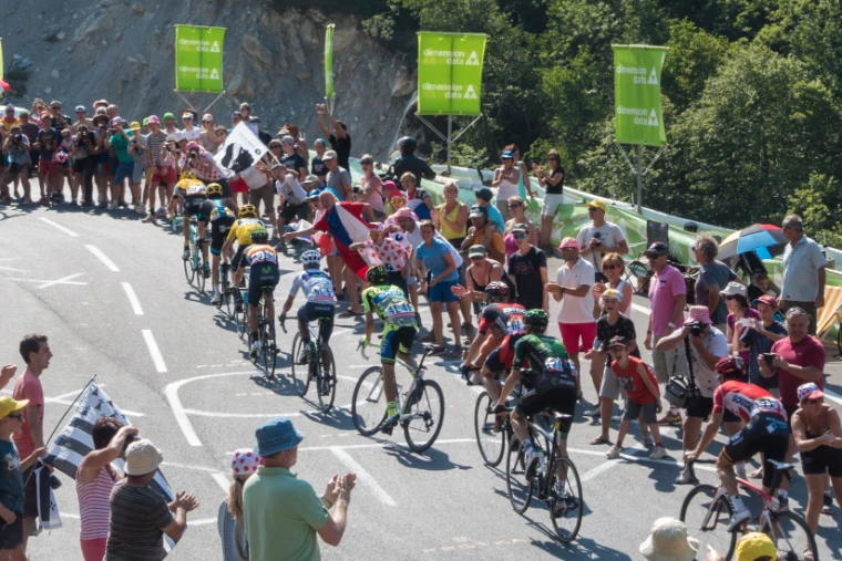Professional riders climb a mountain road through cheering roadside crowds, some waving flags, under green sponsor banners (2015). The Tour de France, Stage 11, Pau to Cauterets, Hautes-Pyrénées, France, in the season calendar of The Mansa Court