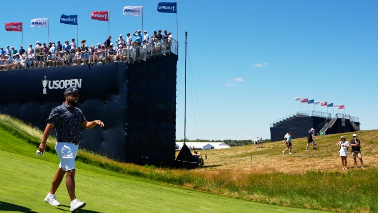 A player walking up the fairway beneath a packed grandstand flying US Open flags, spectators crossing the rough beyond, at the US Open, a mid-June engagement in the season calendar of The Mansa Court