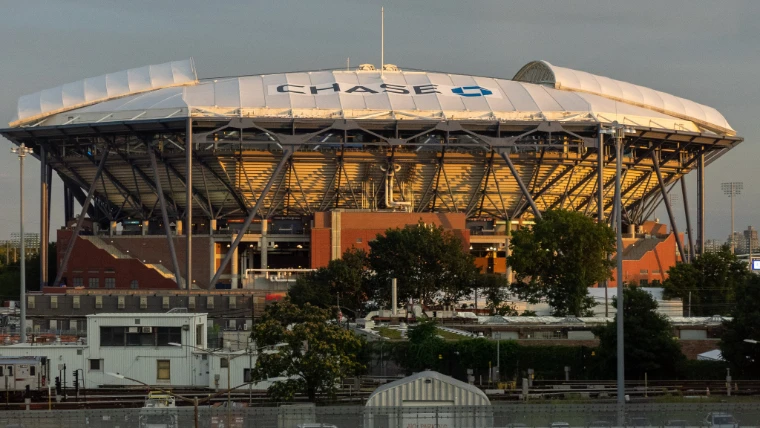 The steel-framed roof of Arthur Ashe Stadium catches warm evening light above a line of green trees (2019).