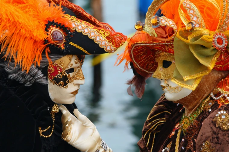 Two carnival masks in gilt and orange plumes turned face to face above a canal at The Venice Carnival, an engagement in the season calendar of The Mansa Court