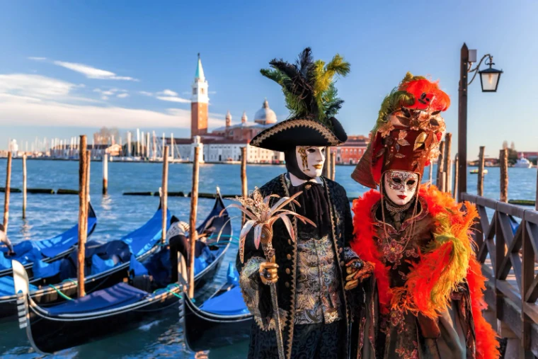 A masked pair in tricorne and plumed headdress on the Riva above the moored gondolas with San Giorgio Maggiore beyond at The Venice Carnival, an engagement in the season calendar of The Mansa Court
