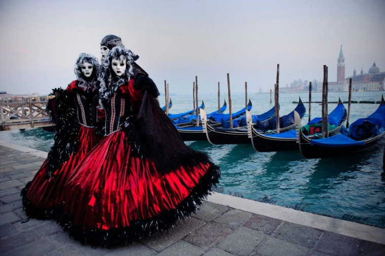Three costumed figures in red and black silk with white masks and powdered wigs beside the moored gondolas at The Venice Carnival, an engagement in the season calendar of The Mansa Court
